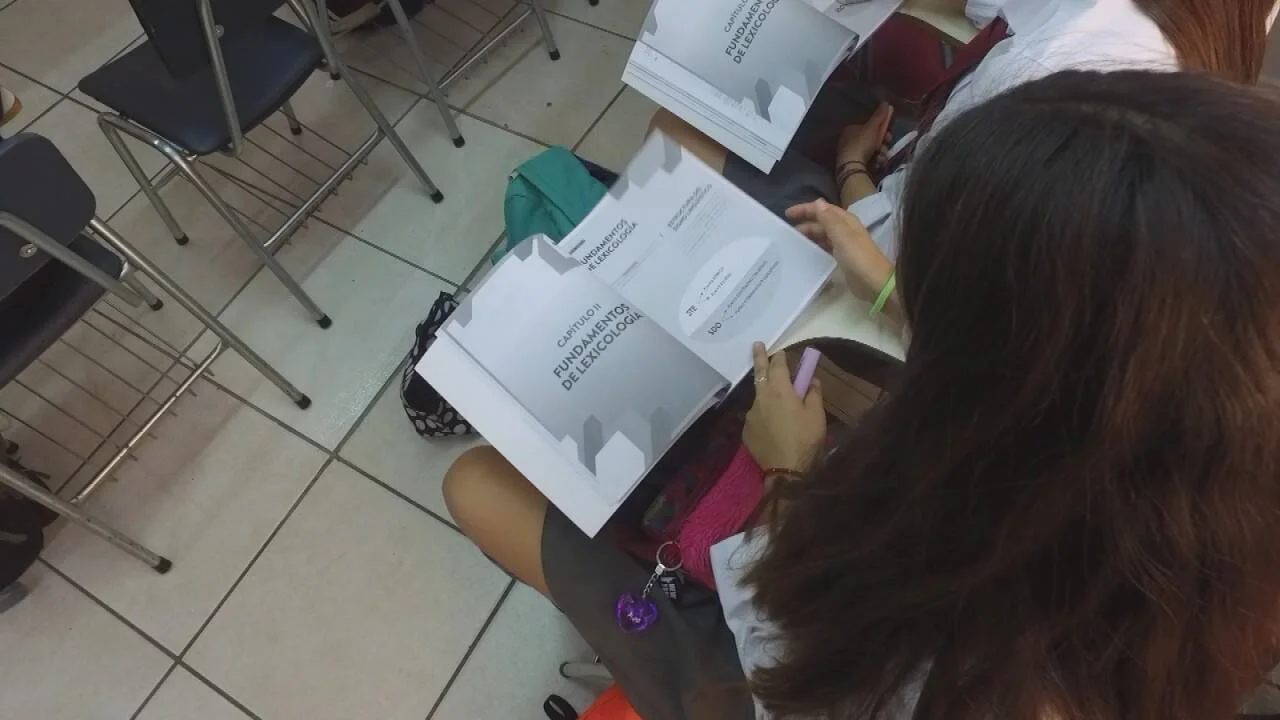 Estudiante sostiene un documento titulado 'Fundamentos de Lexicología' durante una clase universitaria.