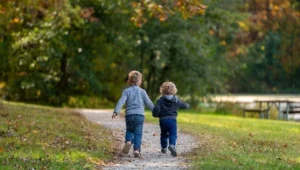 Dos niños pequeños corren tomados de la mano por un sendero de un parque en otoño.