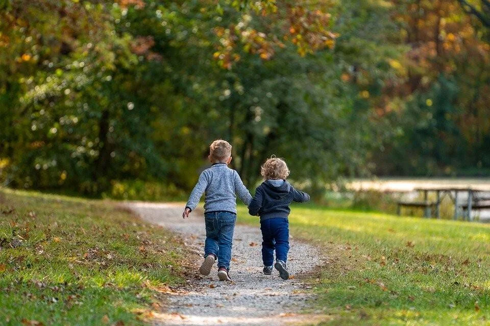 Dos niños pequeños corren tomados de la mano por un sendero de un parque en otoño.