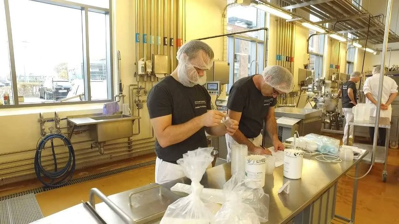 Dos trabajadores con cofias y delantales manipulan envases y bolsas en una planta industrial de procesamiento de alimentos.