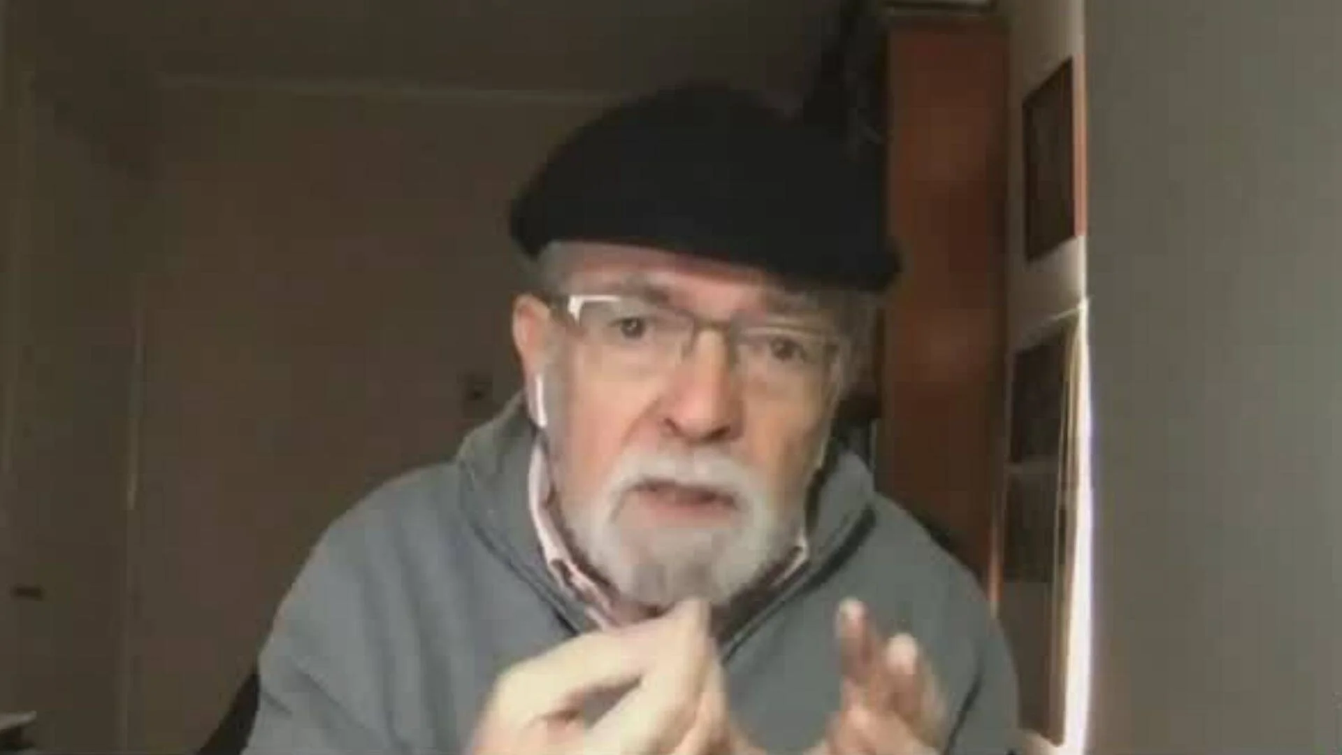 Hombre mayor con boina negra, gafas y barba gris gesticula con las manos mientras habla, en un entorno doméstico.