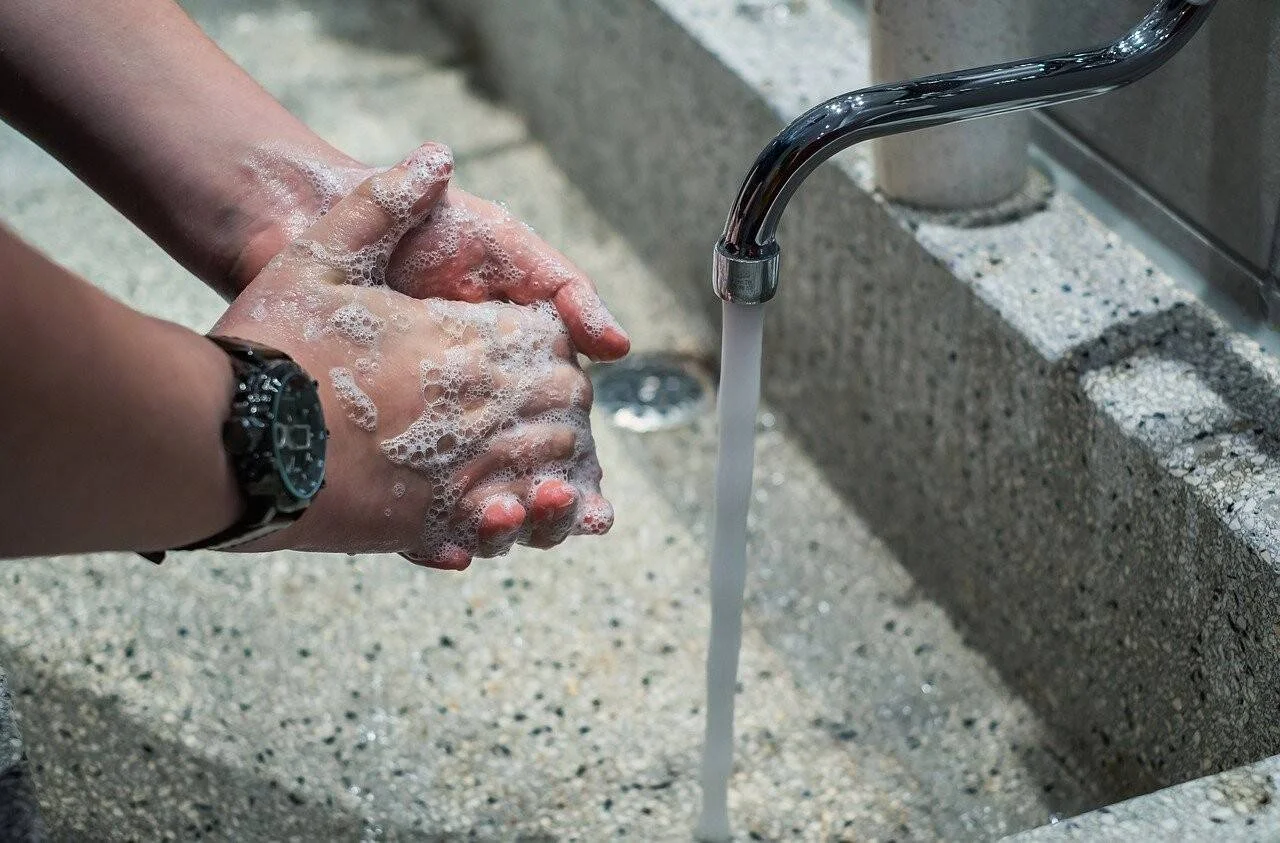 Persona enjabonándose las manos con espuma bajo el chorro de agua de un grifo metálico, con reloj en la muñeca.