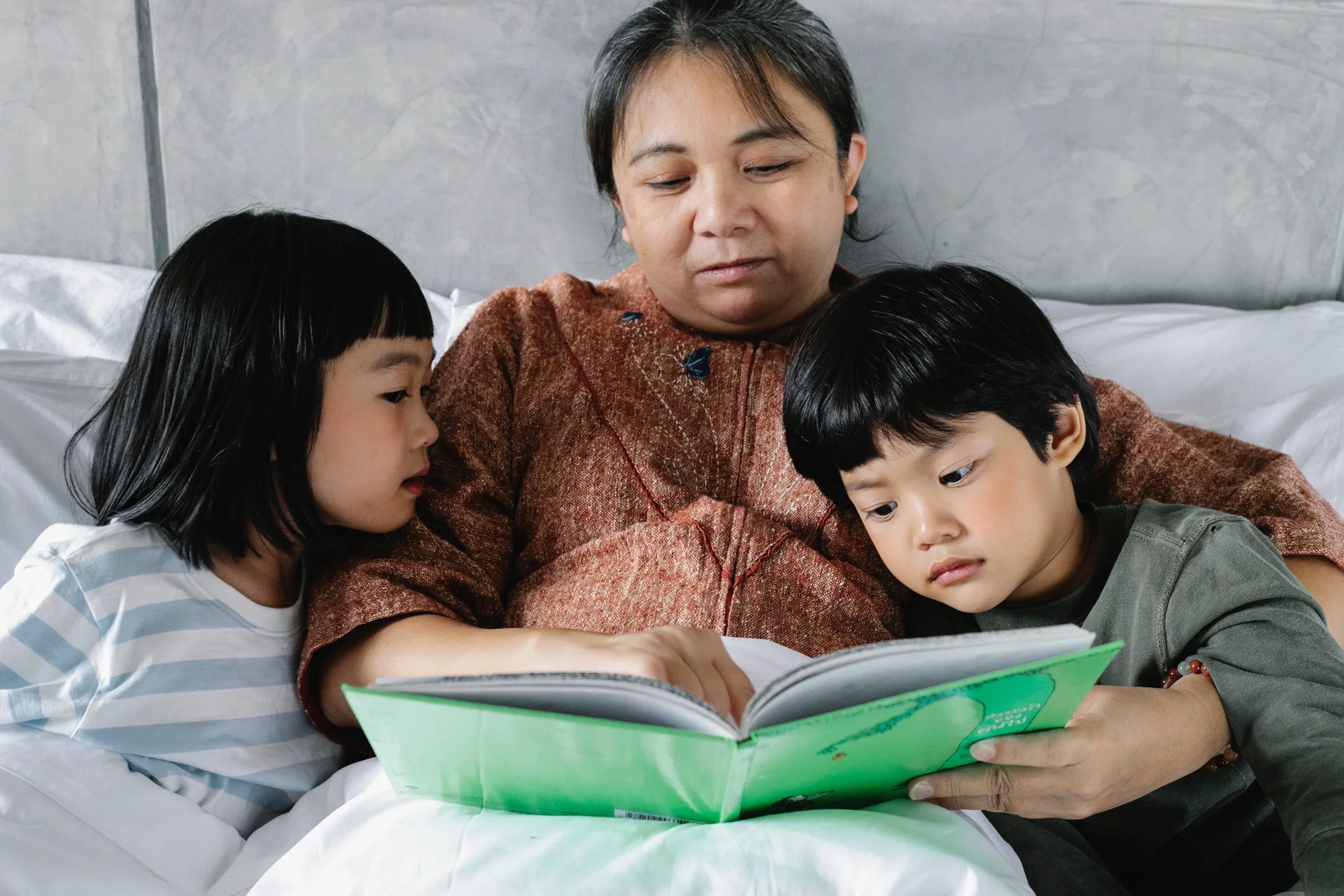 Mujer adulta lee un libro infantil de tapa verde a dos niños pequeños recostados junto a ella en una cama.