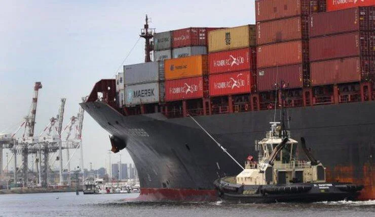 Un gran buque portacontenedores cargado con contenedores de Maersk y Hamburg Süd es guiado por un remolcador en un puerto industrial.