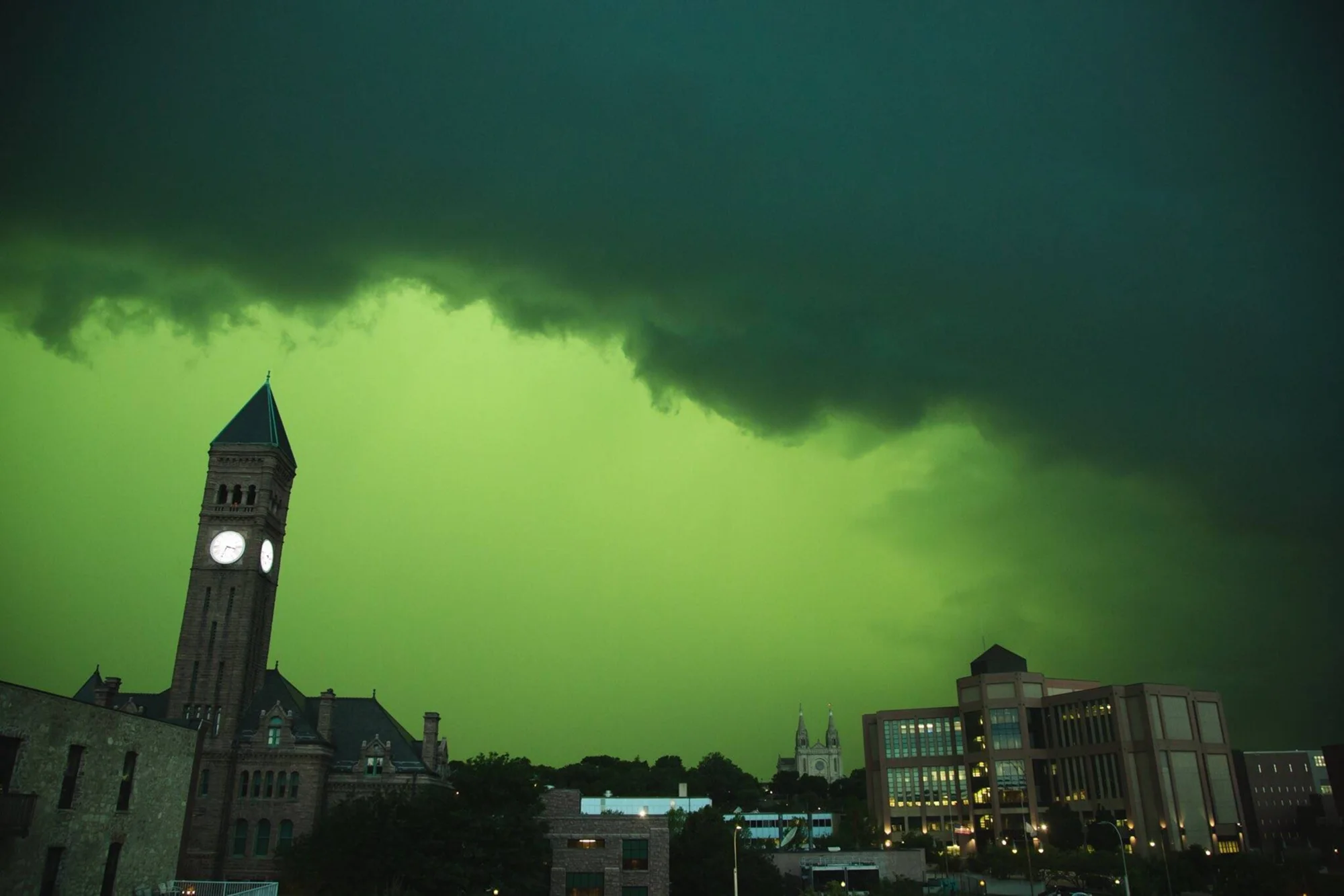 Nubes de tormenta severa tiñen el cielo de verde intenso sobre una ciudad con torre de reloj histórica y edificios modernos.