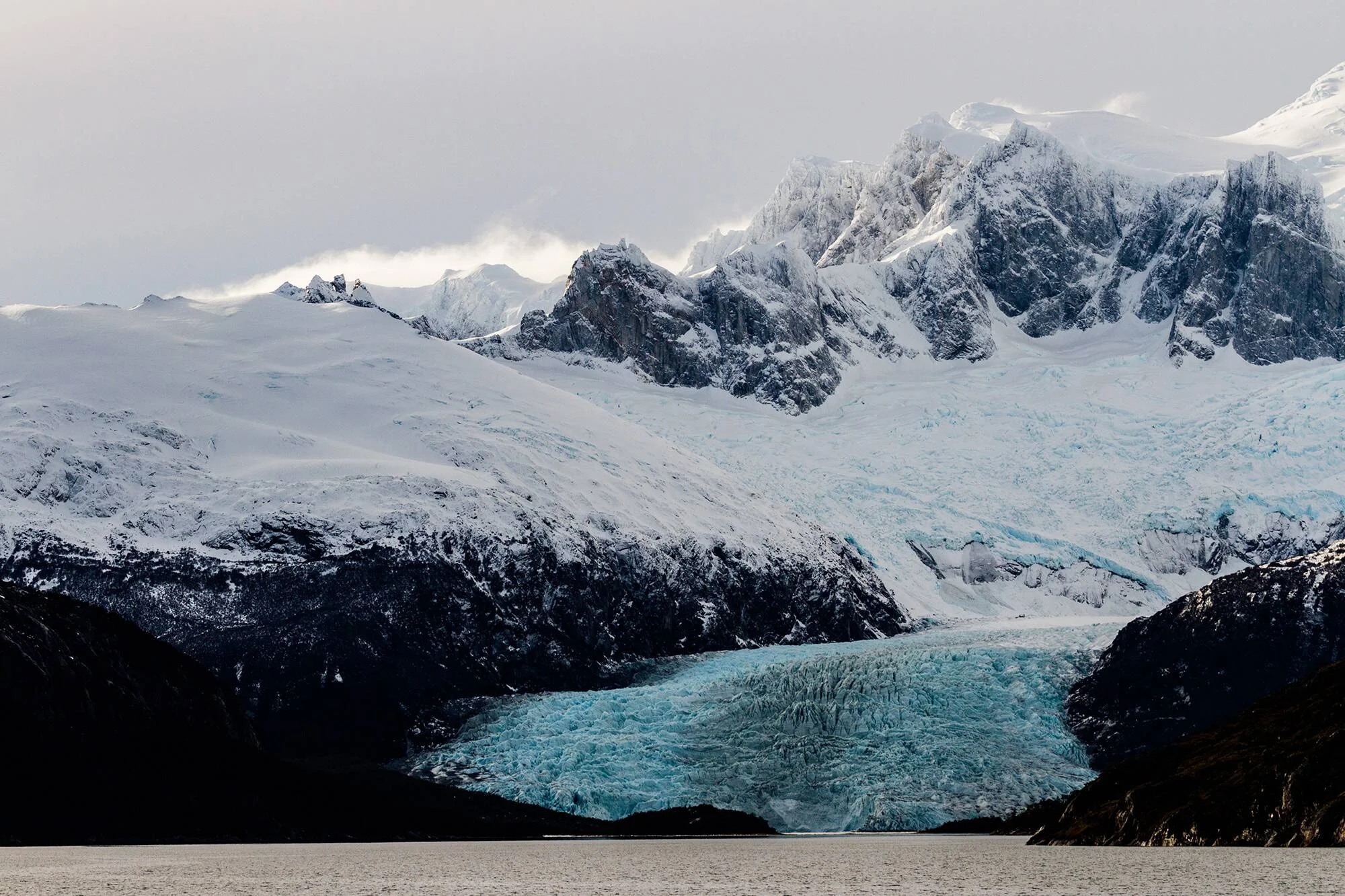 Glaciar de hielo azul desciende por montañas nevadas hasta las aguas de un lago en la Patagonia.