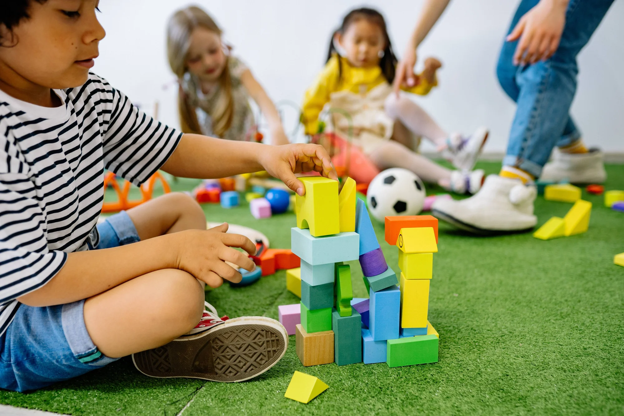 Niño construye una torre con bloques de madera de colores mientras otros niños juegan en el suelo de una sala de juegos.