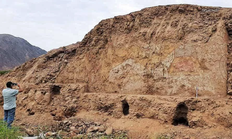 Una persona fotografía un gran muro de adobe con relieves pintados de una figura prehispánica, con nichos excavados en la base.