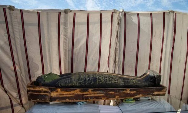 Sarcófago egipcio pintado de negro con inscripciones doradas, expuesto sobre una base de madera en un sitio arqueológico.