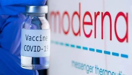 Mano sosteniendo un frasco de vacuna contra COVID-19 frente al logotipo de la farmacéutica Moderna.