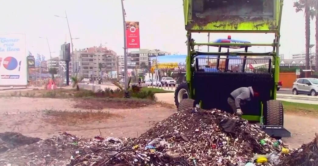 Un trabajador opera una máquina recolectora verde junto a un gran montículo de basura en una avenida costera de una ciudad.