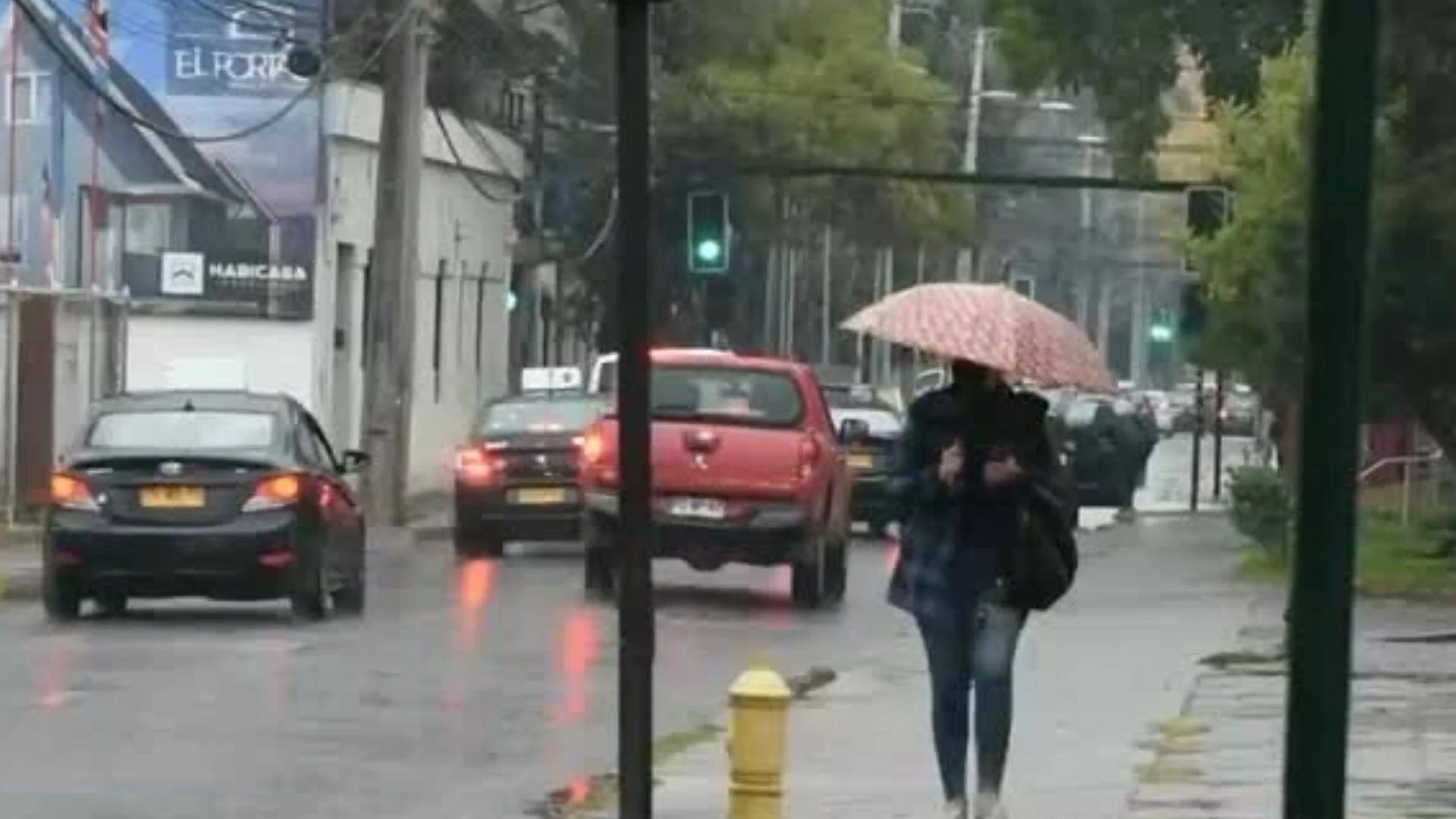Una persona camina con paraguas por una vereda mojada mientras autos circulan por la calle bajo la lluvia.