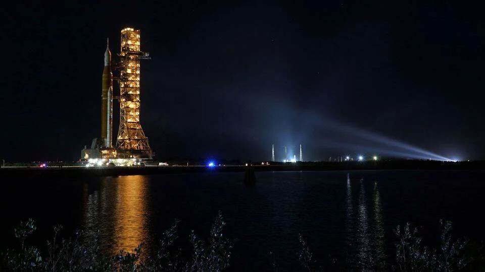 Gran cohete espacial sobre su plataforma de lanzamiento, iluminado de noche y reflejado en el agua cercana.