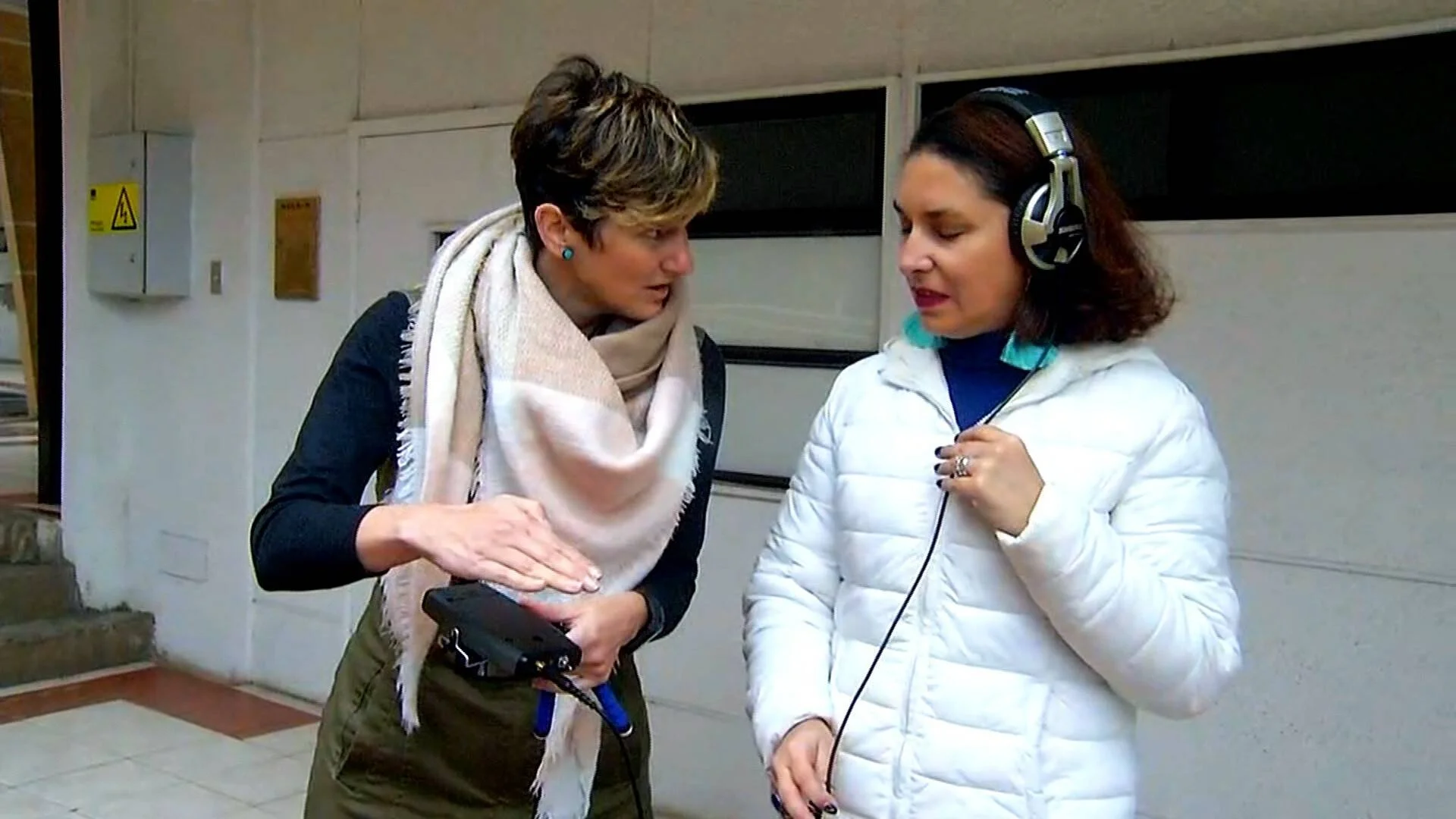 Una mujer opera una grabadora portátil mientras otra, con auriculares, escucha atentamente en un espacio exterior.