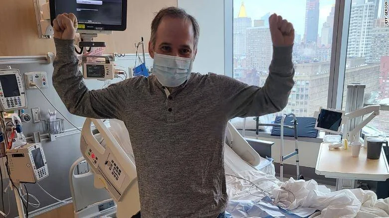 Hombre con mascarilla levanta los puños en señal de victoria en una habitación de hospital con vista a una ciudad.
