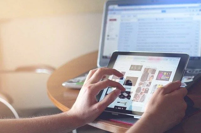 Manos de una persona navegando por contenido en una tablet, con una computadora de escritorio encendida en el fondo.
