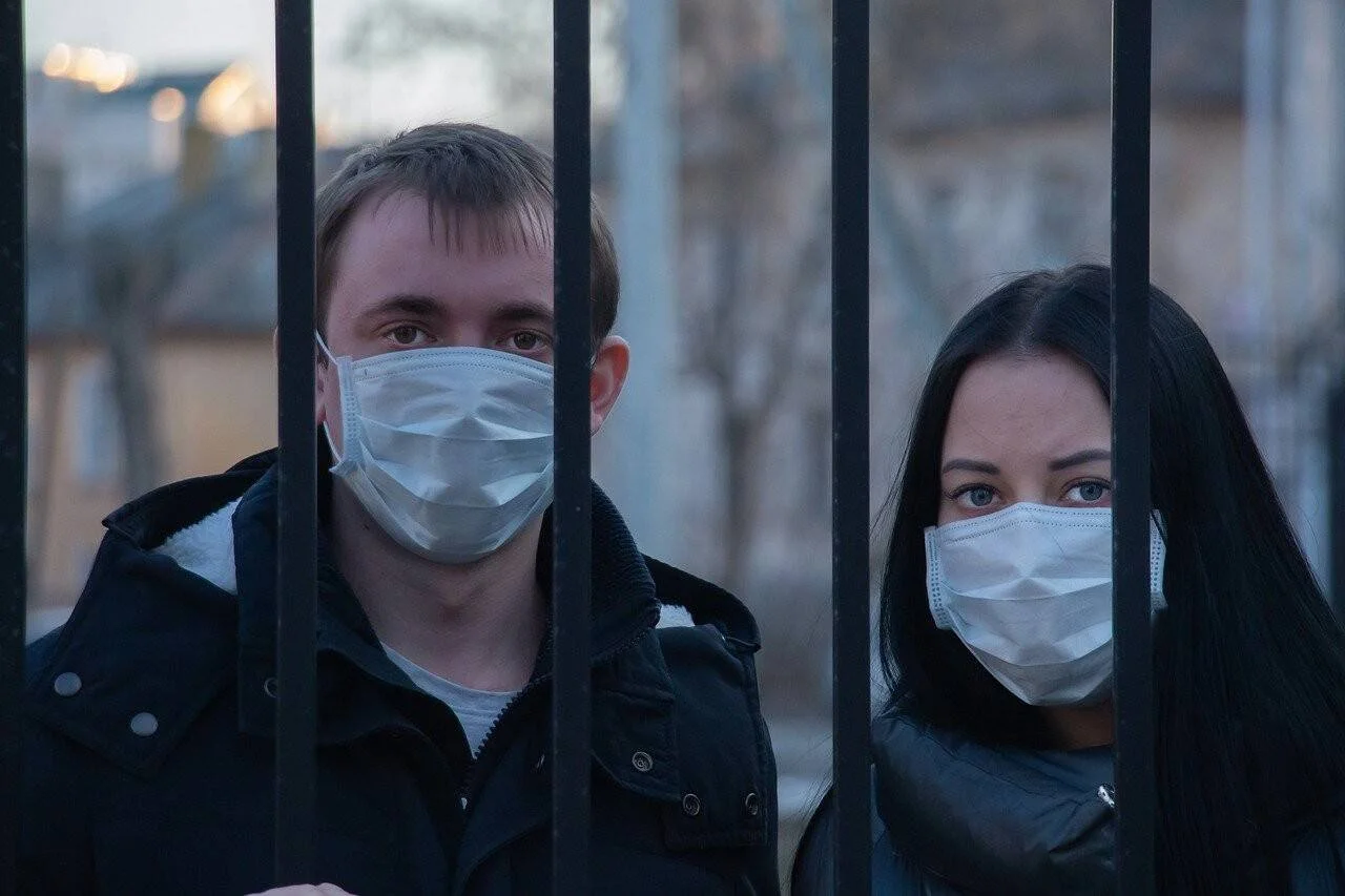 Un hombre y una mujer con mascarillas quirúrgicas miran a cámara desde detrás de una reja metálica en un entorno urbano.