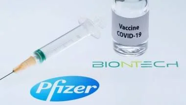 Frasco de vacuna COVID-19 junto a una jeringa sobre logos de Pfizer y BioNTech.