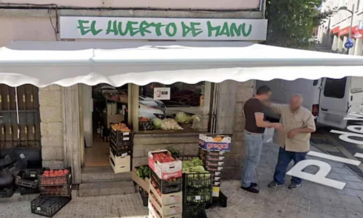 Fachada de la frutería 'El Huerto de Manu' con cajas de frutas y verduras en la acera y dos personas conversando.