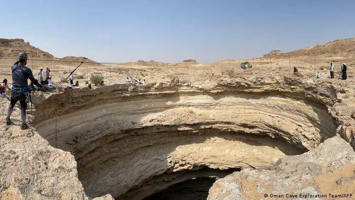 Un escalador equipado se asoma al borde de una enorme cueva en paisaje desértico rocoso, rodeado de personas observando.