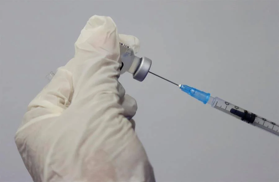 Mano con guante blanco sostiene un frasco de medicamento mientras inserta una jeringa para extraer una dosis.