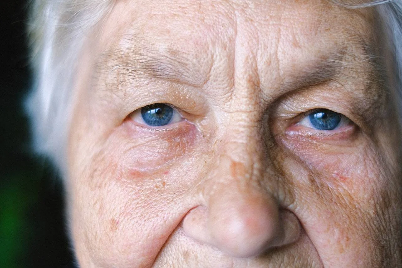 Primer plano del rostro de una mujer mayor de cabello blanco y ojos azules, con arrugas marcadas y expresión seria.
