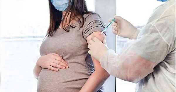 Mujer embarazada con mascarilla recibe una inyección en el brazo de un profesional de salud con traje protector.