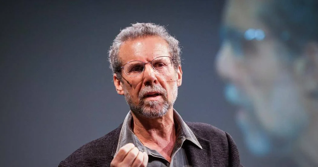 Hombre mayor con gafas y barba canosa habla con gesto enfático ante un auditorio, con una proyección difusa de fondo.