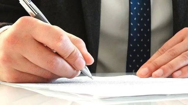 Persona con traje y corbata firma un documento con bolígrafo sobre una mesa.