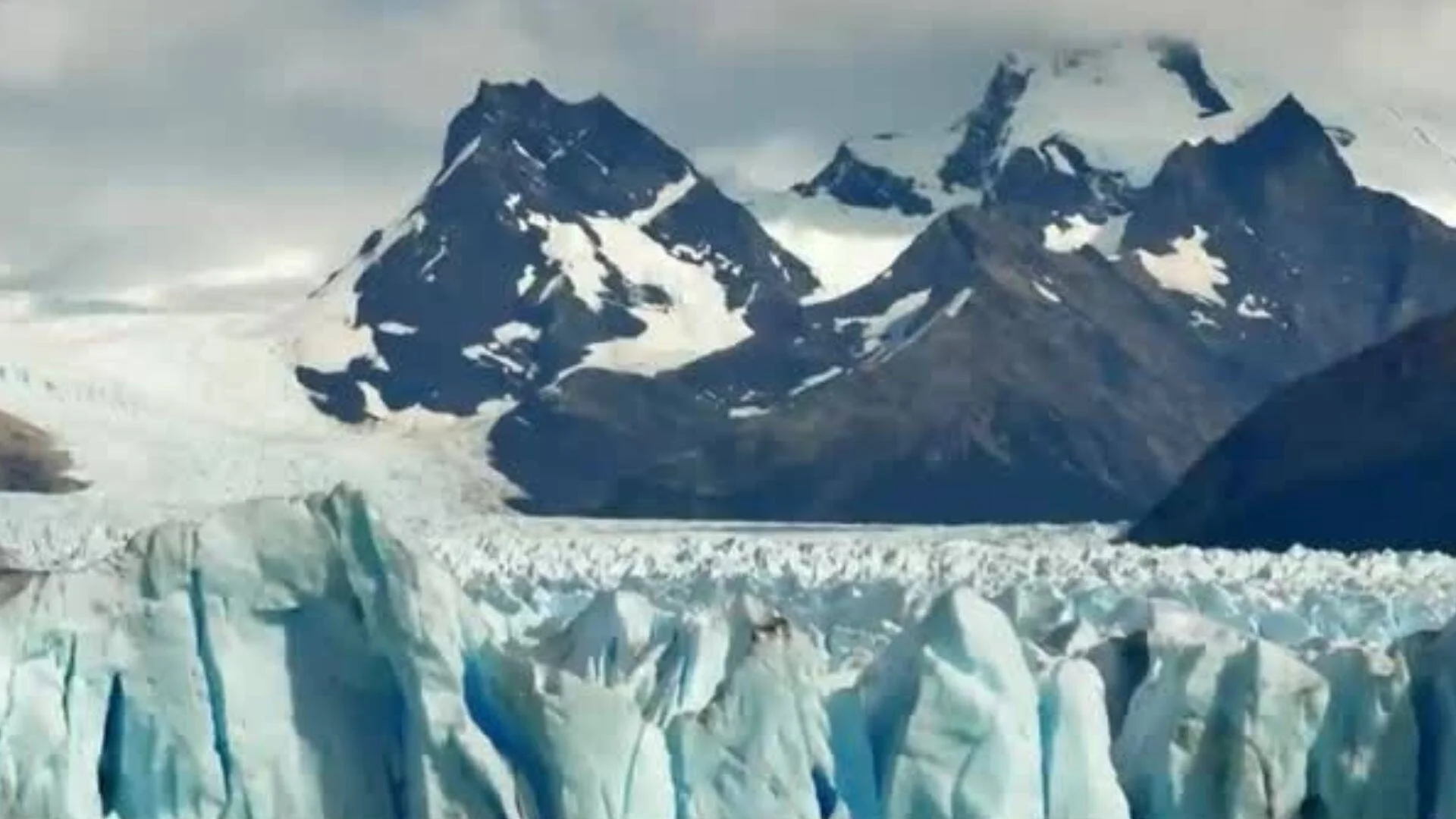 Pared frontal de un glaciar con hielo azul en primer plano y montañas nevadas al fondo bajo cielo nublado.