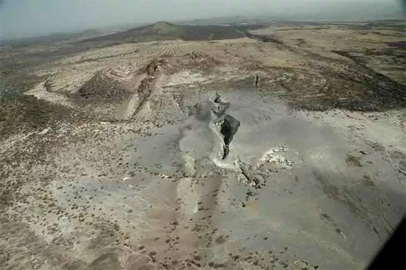 Vista aérea de un volcán de barro activo en un paisaje desértico y árido, con una columna de lodo emergiendo del cráter.