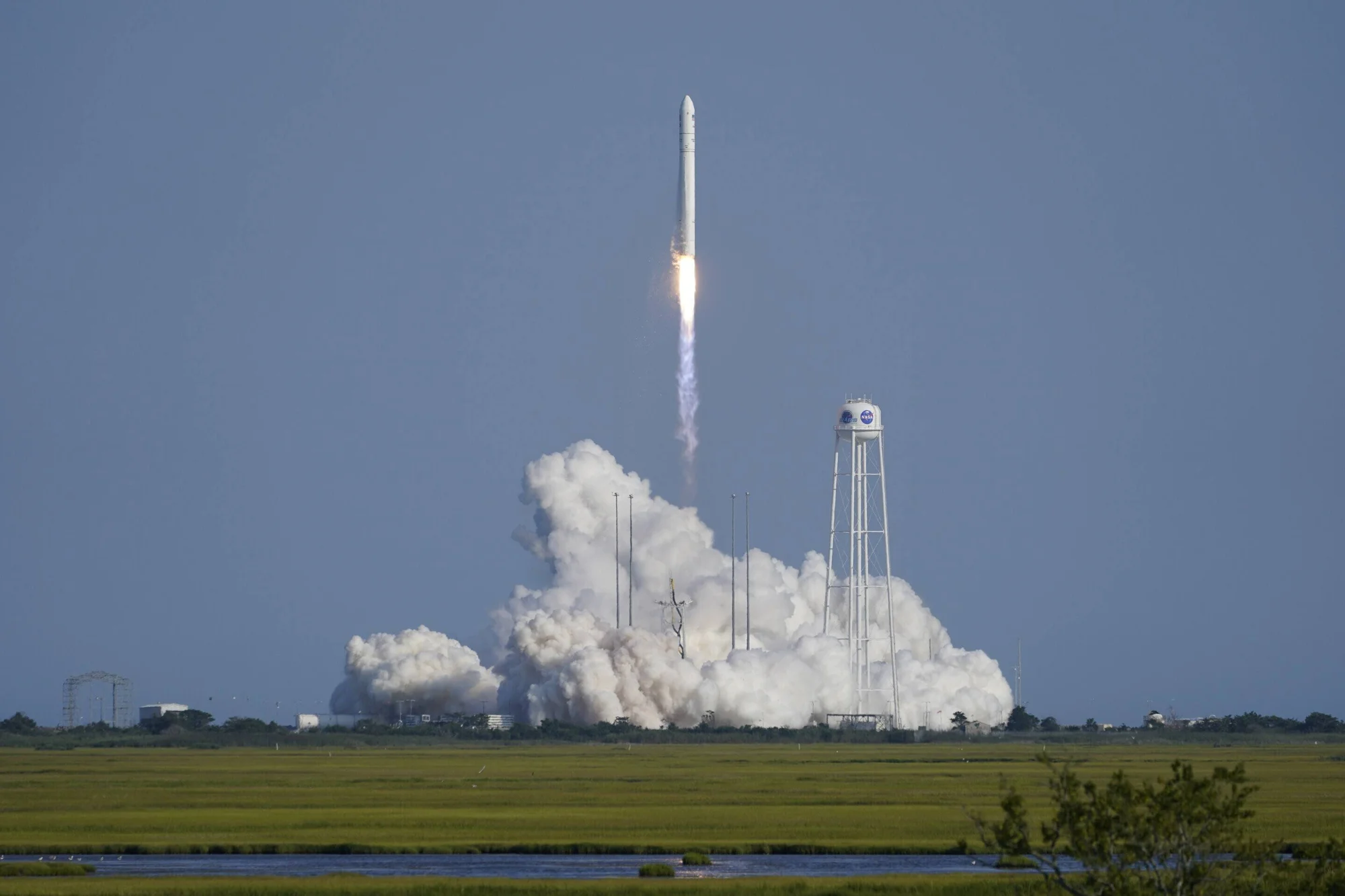 Un cohete despega con una gran nube de humo blanco junto a una torre de agua con el logo de la NASA, en una zona costera de vegetación baja.