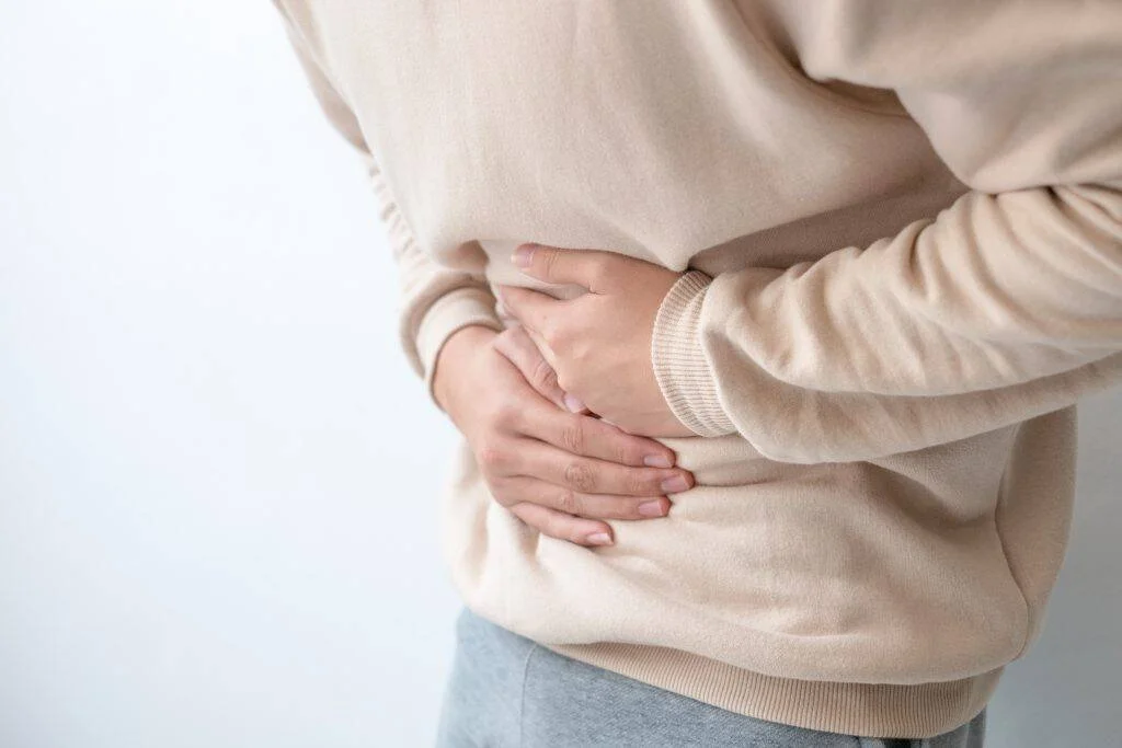 Persona con suéter beige sostiene su abdomen con ambas manos, indicando dolor o malestar estomacal.
