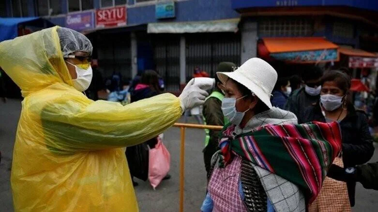 Trabajador de salud con traje amarillo toma la temperatura a una mujer con tapabocas en una calle concurrida.