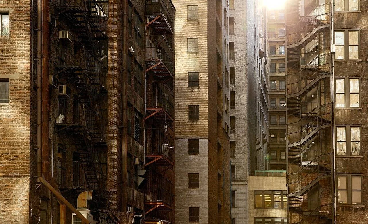 Callejón urbano flanqueado por edificios de ladrillo con escaleras de incendio metálicas y luz solar entre las fachadas.