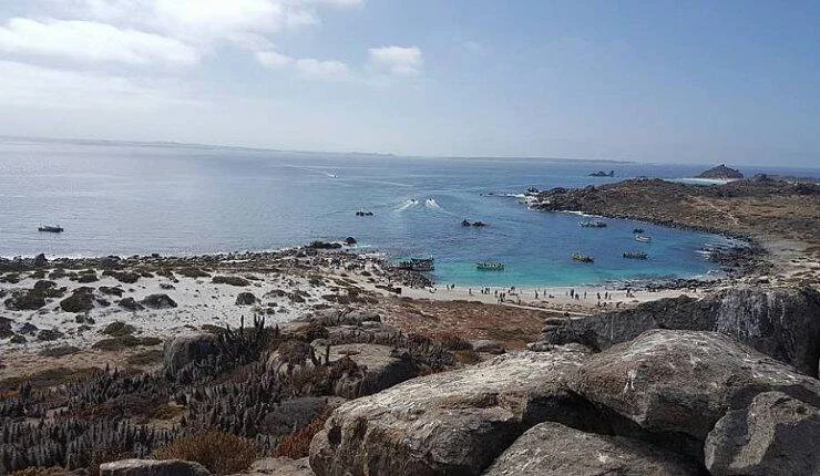 Vista panorámica de una bahía de aguas turquesas con embarcaciones y playa rodeada de rocas y vegetación árida.
