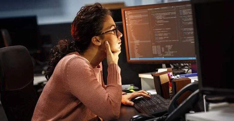 Mujer con gafas observa concentrada múltiples monitores con código de programación en oficina oscura.