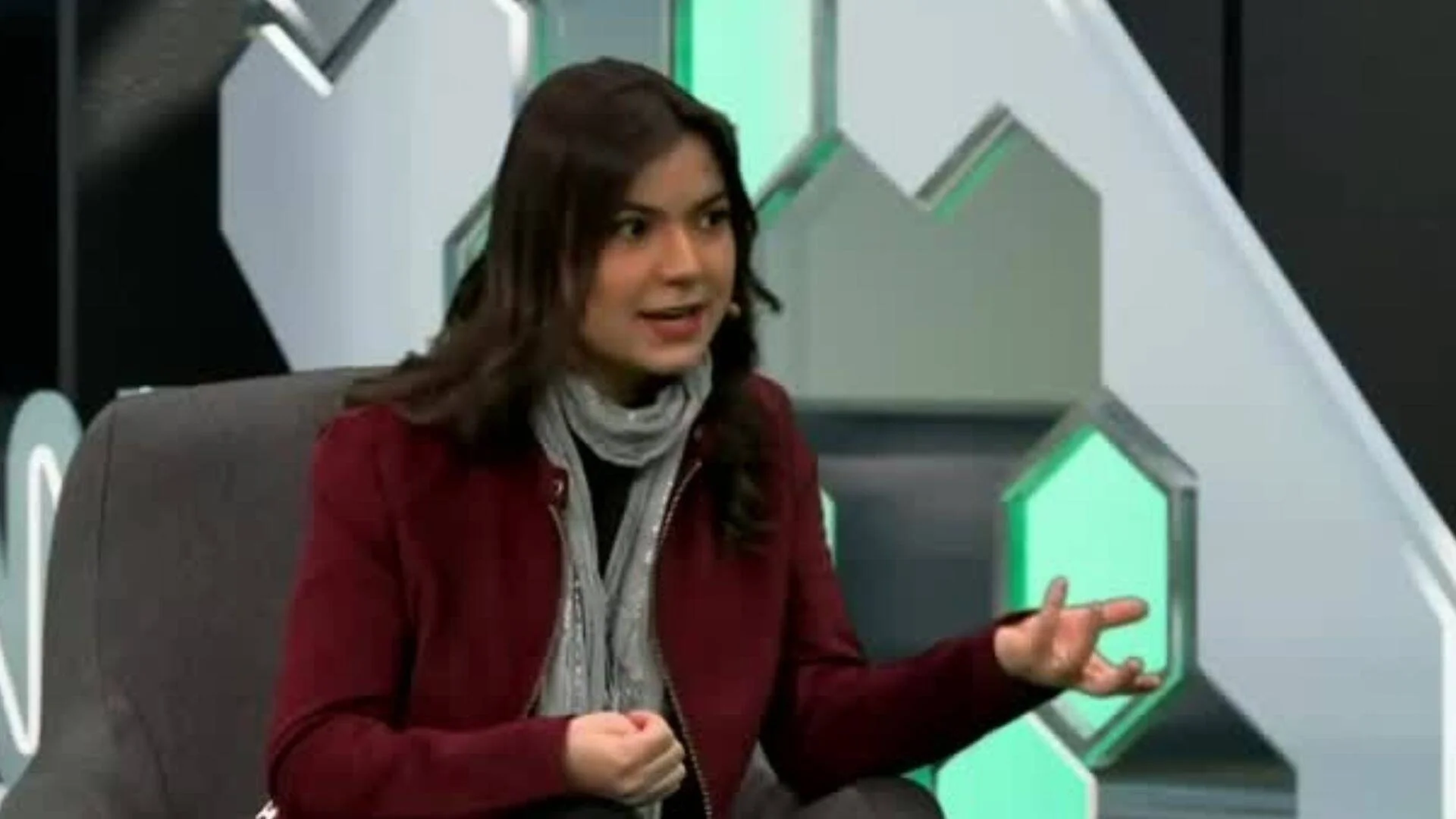 Mujer joven con saco bordo y bufanda gris habla gesticulando en un estudio de televisión con decorado geométrico verde.