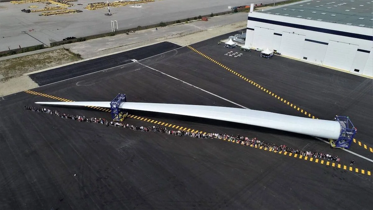 Vista aérea de una enorme pala de turbina eólica apoyada en soportes, flanqueada por cientos de personas que ilustran su escala colosal.