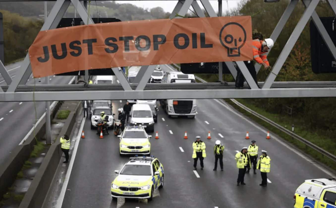 Activista cuelga una pancarta naranja de 'Just Stop Oil' en un puente mientras la policía corta el tráfico en la autopista.