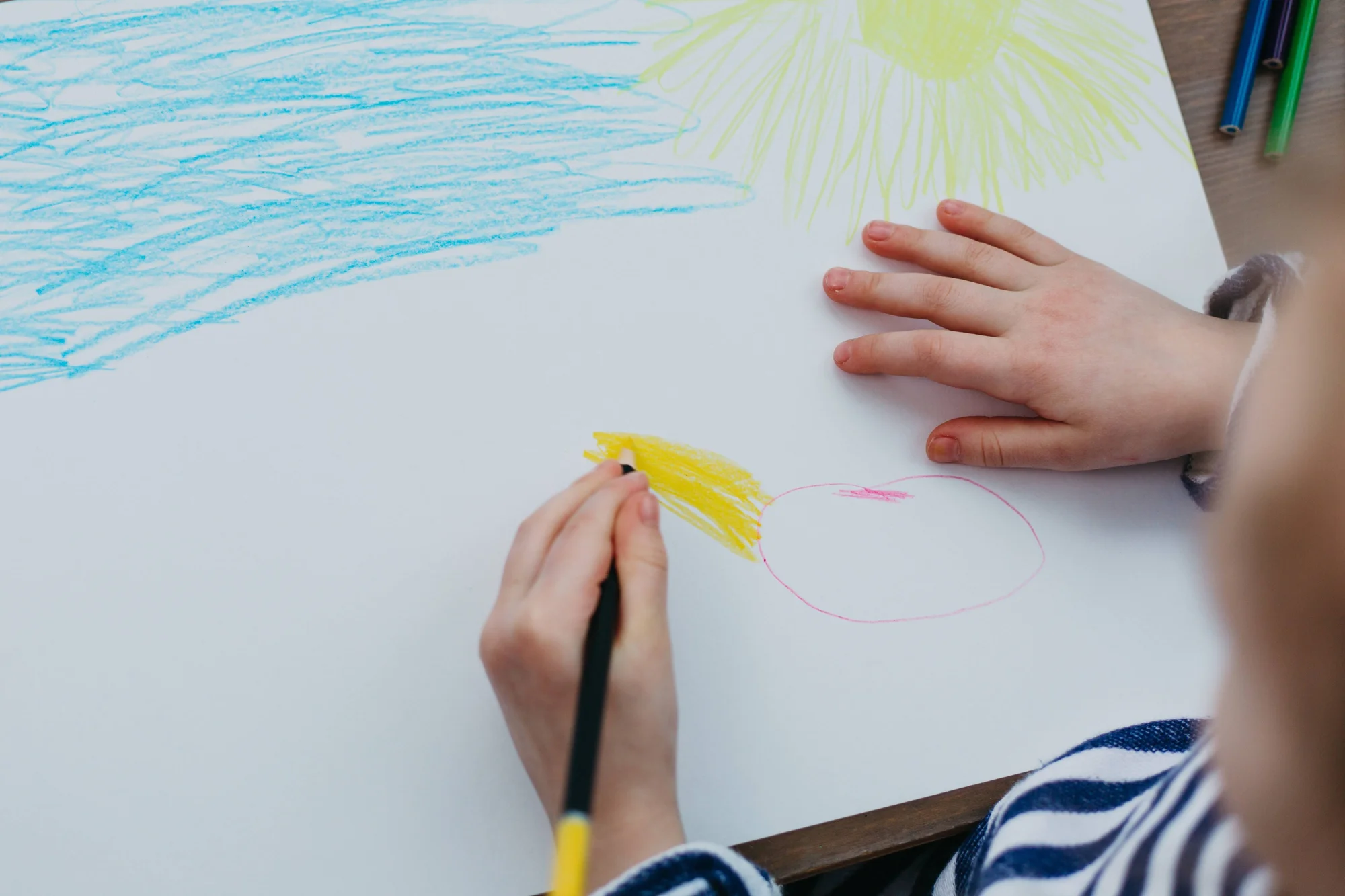 Manos de un niño coloreando un dibujo con lápices de colores sobre papel blanco, con figuras de cielo y sol.