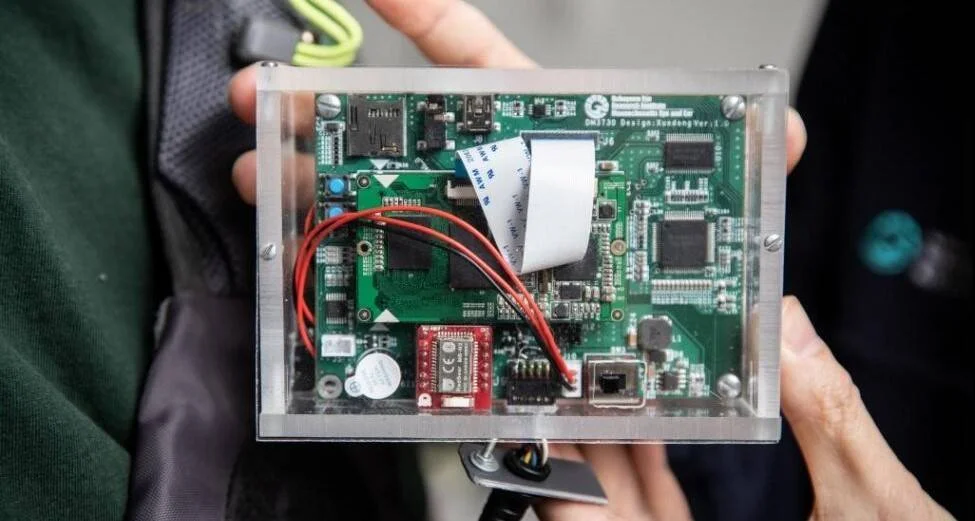 Manos sosteniendo una caja acrílica transparente que contiene una placa de circuito impreso verde con cables y componentes electrónicos.