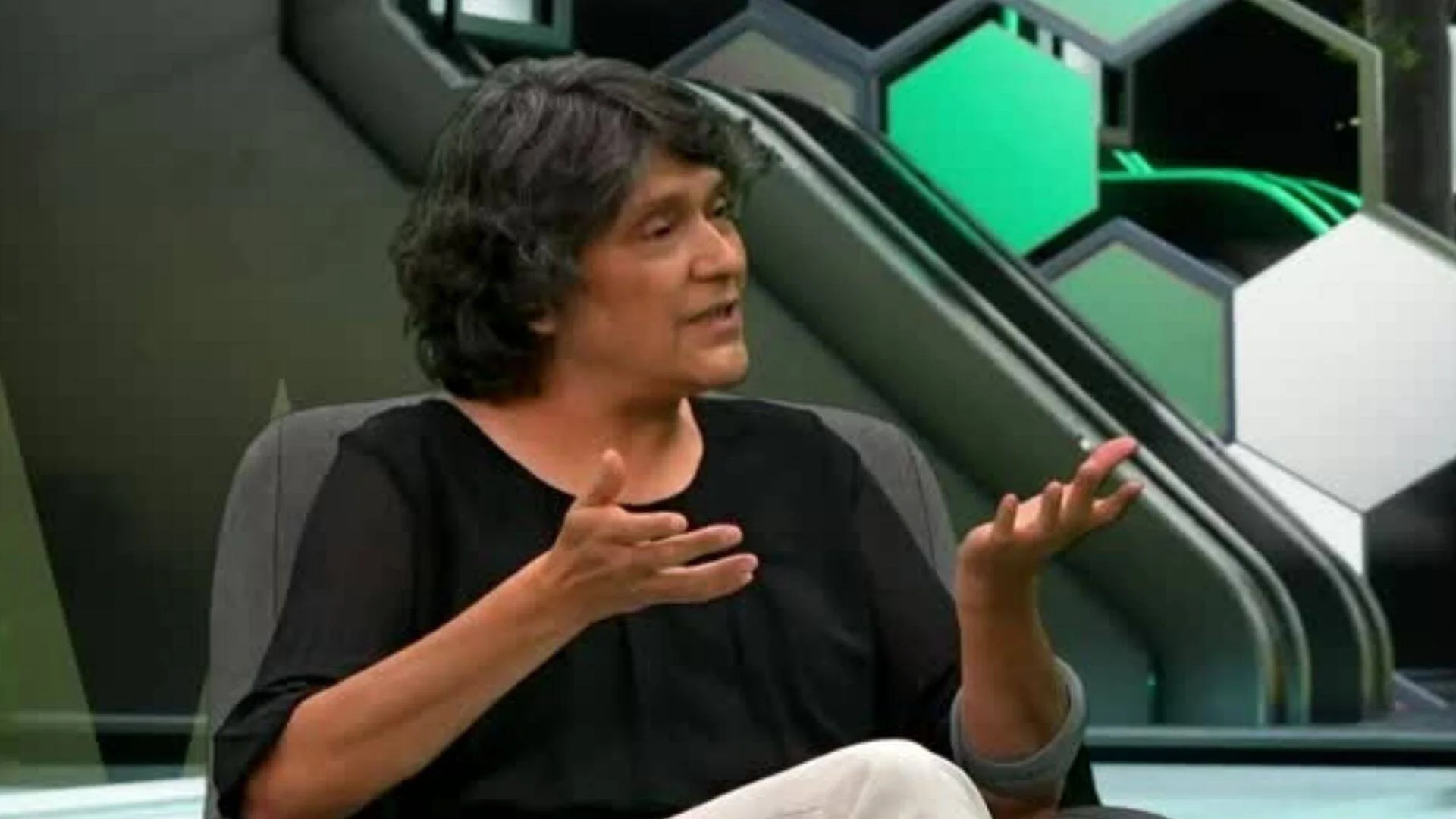 Mujer de cabello oscuro con remera negra gesticula mientras habla sentada en un estudio de televisión con decorado geométrico verde.