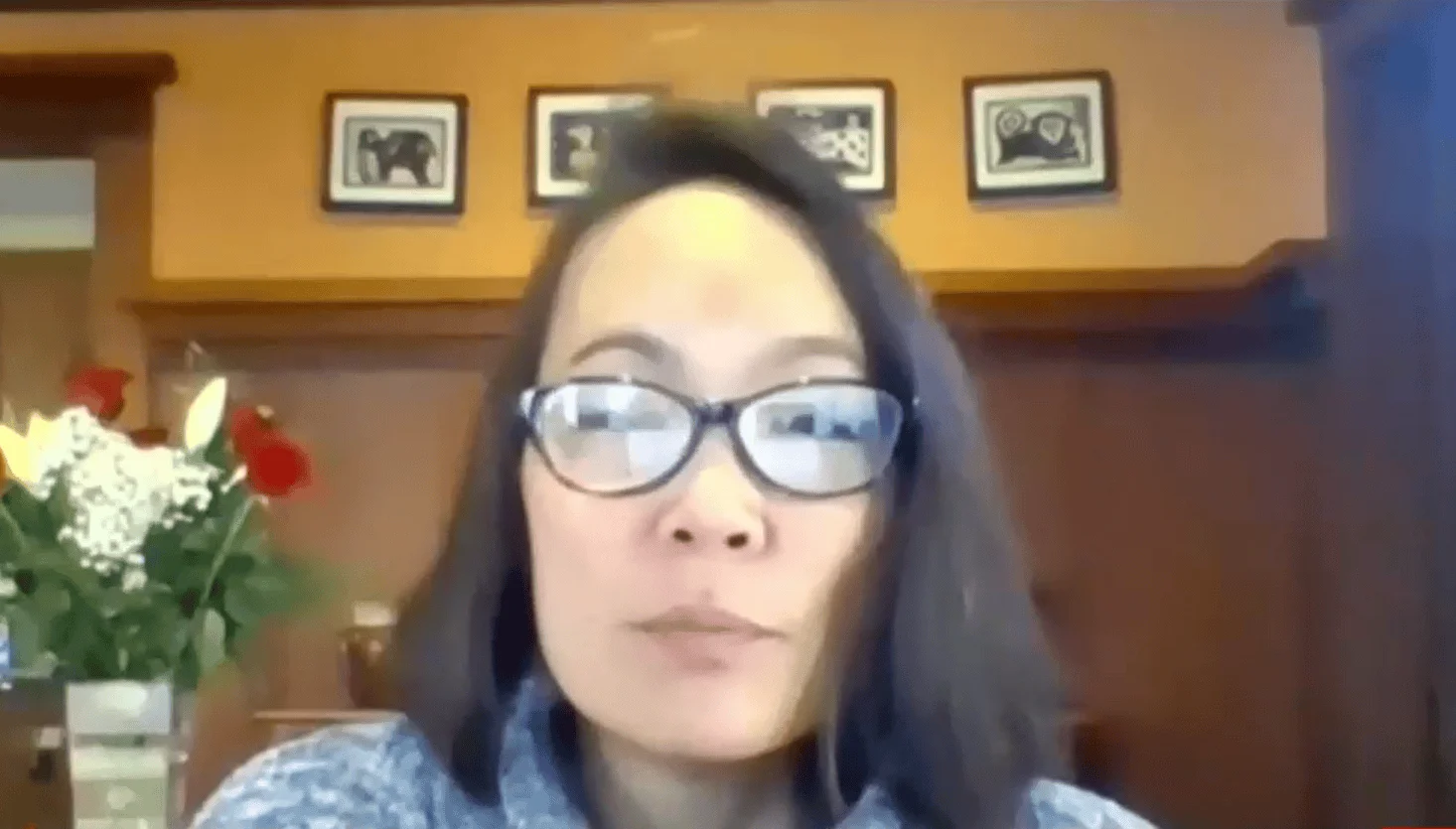 Mujer de cabello oscuro y anteojos participa en una videollamada desde una oficina con cuadros y flores al fondo.