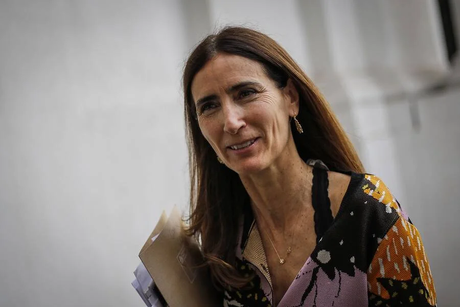 Mujer de cabello castaño largo sonríe mientras camina con documentos en la mano, vistiendo blusa estampada colorida.