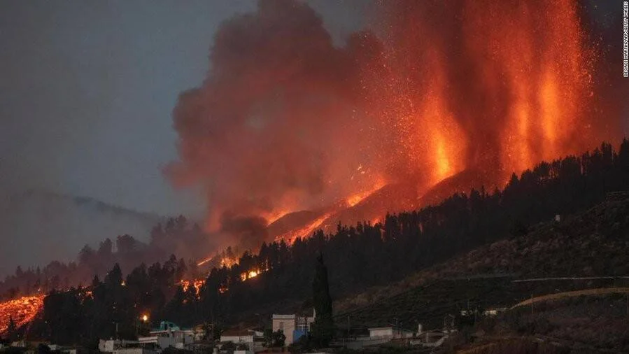 Volcán en erupción expulsa lava y ceniza sobre una ladera arbolada mientras flujos ígneos descienden hacia viviendas.
