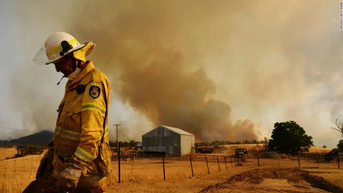 Bombero con equipo de protección camina frente a una granja mientras enormes columnas de humo se elevan por un incendio forestal.