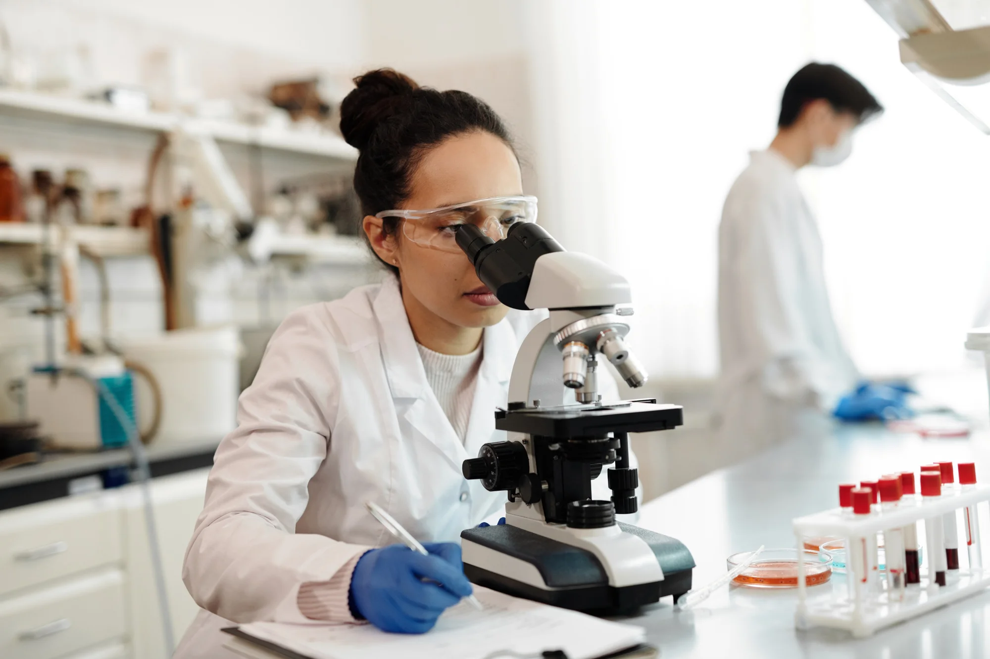 Investigadora con bata blanca y guantes azules observa muestras al microscopio mientras toma notas en un laboratorio.