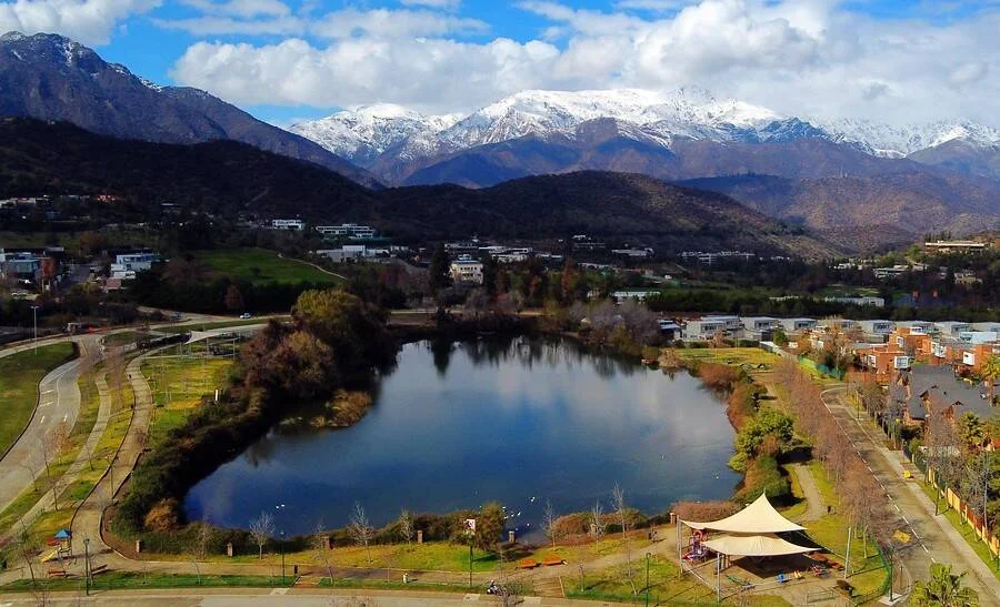 Vista aérea de una laguna en parque urbano rodeada de edificaciones, con la Cordillera de los Andes nevada al fondo.