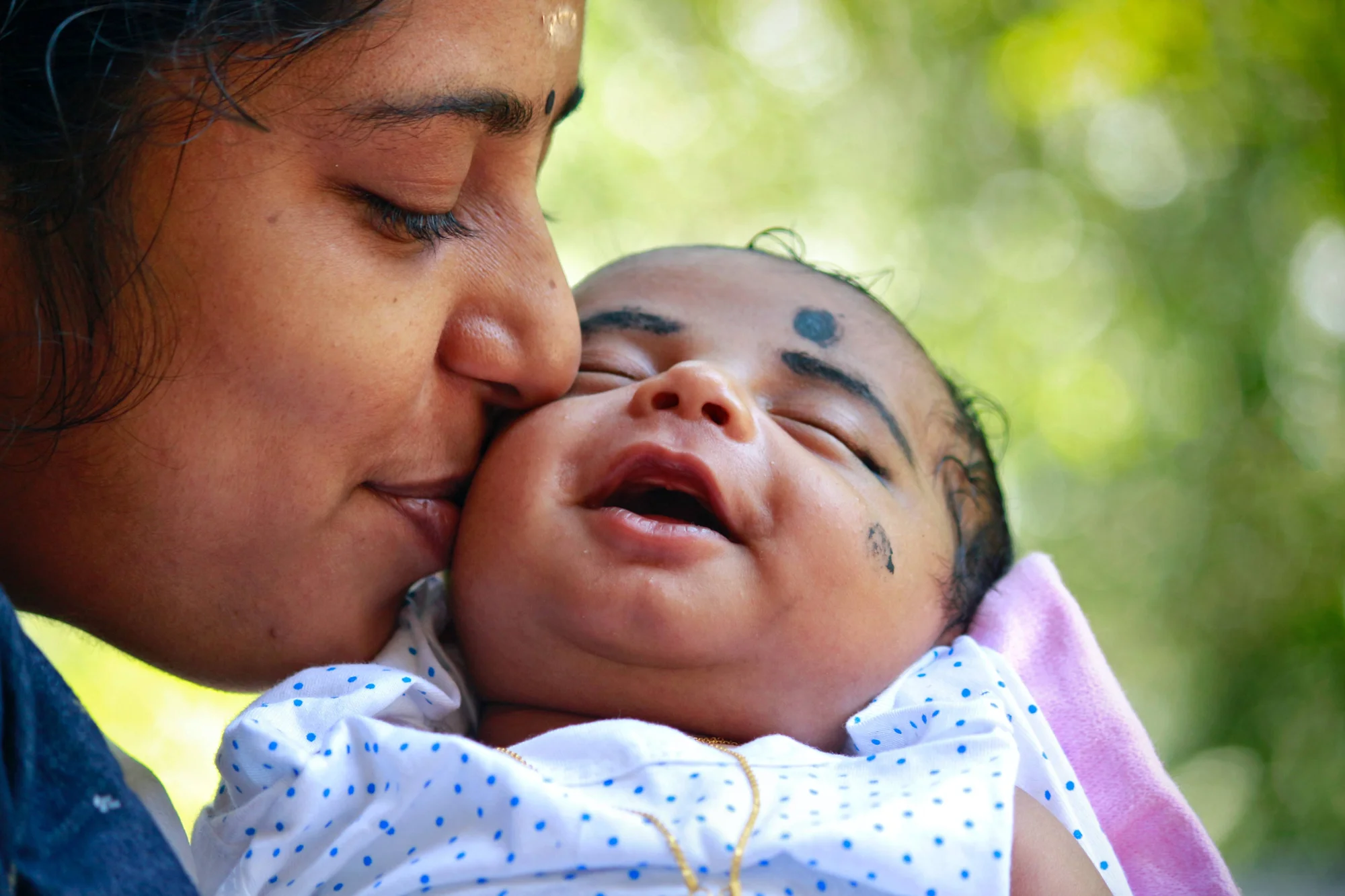 Mujer india besa tiernamente la mejilla de su bebé recién nacido con bindi en la frente, al aire libre.